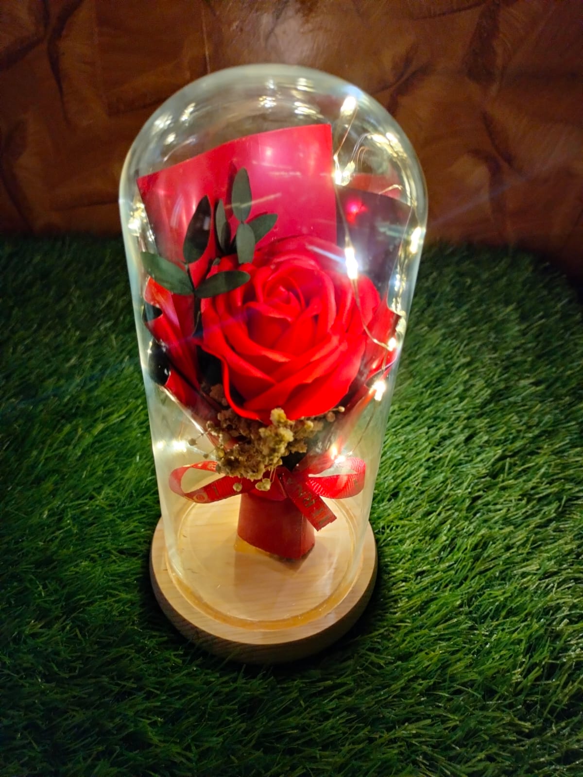 Decorative flower arrangement in a glass dome with LED lights and ribbon on wooden base.